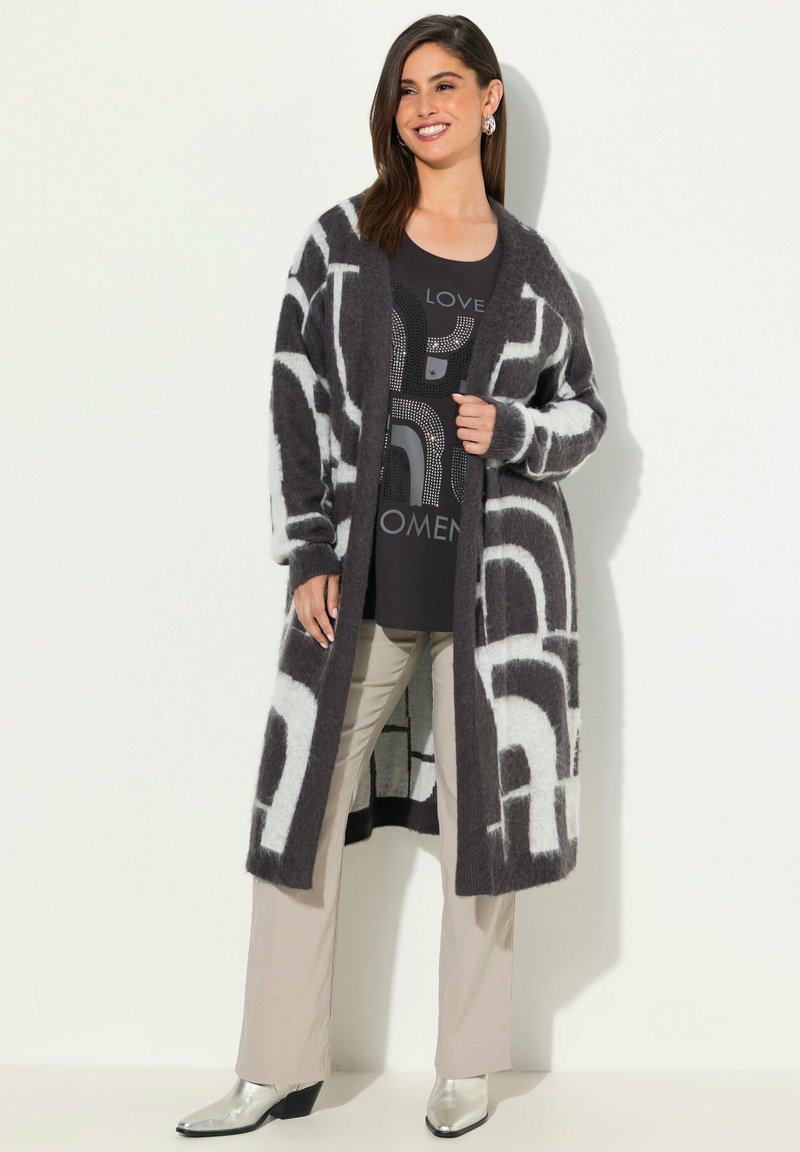 Langer, schwarz-weißer Cardigan mit Muster, aus flauschigem Material, in lockerer Passform. Kombiniert mit einem dunklen Shirt, hellen Hosen und silbernen Stiefeln.
