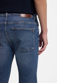 Tmavomodré denimové džínsy s koženou značkou loga na zadnej strane, dvomi zadnými vreckami a detailmi stehu viditeľnými na látke.