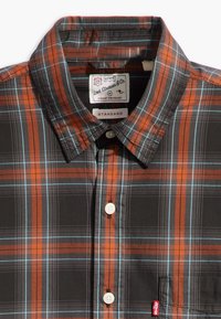Camisa de cuadros en negro, naranja y azul. Presenta un cierre de botones en la parte delantera, cuello tipo camisa y un bolsillo en el pecho con una etiqueta de logo.