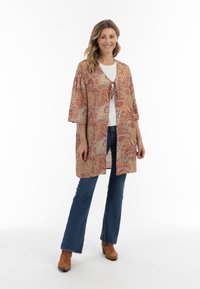 Cardigan in stile kimono con motivo a paisley in rosso, oro e blu, con maniche a tre quarti e un nodo al collo, indossato sopra un top bianco.