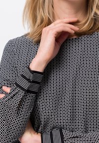 Eine Frau mit blonden Haaren trägt eine schwarz-weiß gemusterte Bluse mit langen Ärmeln und gestreiften Manschetten. Ihre Hand ruht nahe am Kinn, während sie die Arme verschränkt.