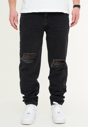Black denim jogger pants featuring distressed knee areas, a relaxed fit, and elastic cuffs, paired with white sneaker shoes.