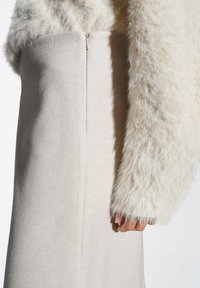 Gros plan sur une personne portant un pull en crème duveteux avec une jupe beige taille haute présentant une fermeture éclair visible au dos.