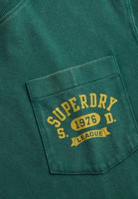 Gros plan sur un tissu bleu sarcelle présentant une poche poitrine avec un texte jaune : "SUPERDRY S. 1976 D. LEAGUE" sur un vêtement en coton.