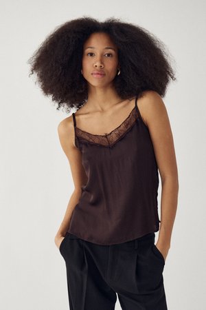 Giovane donna con afro naturale che indossa un top color marrone scuro con pizzo e pantaloni neri, in piedi con le mani nelle tasche.