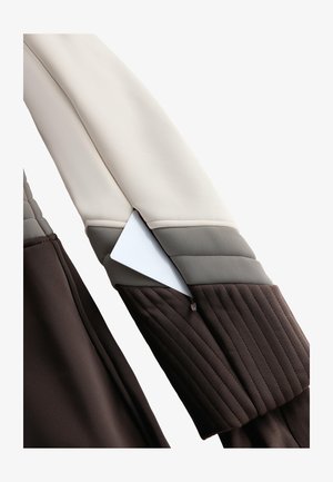 Bruin en beige jassmouw met gestructureerd stiksel, voorzien van een witte kaartenzak. Zachte, gladde stof met kleurvlakken ontwerp.