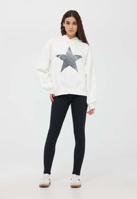 Felpa bianca con una grande grafica a stella grigia luccicante sulla parte anteriore, abbinata a leggings neri e sneakers bianche con suole marroni.