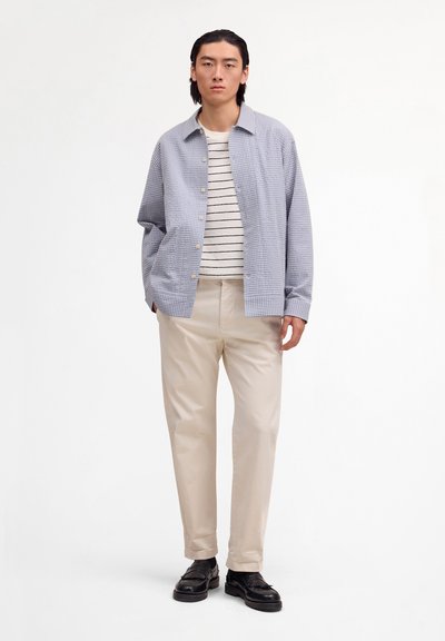 Jeune homme portant une veste à carreaux bleu clair, une chemise rayée blanche et noire, un pantalon beige et des mocassins en cuir noir sur fond blanc.