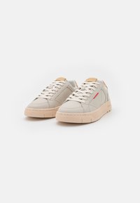 Sneaker in pelle beige con una finitura testurizzata, caratterizzati da un accento contrastante color nocciola, lacci bianchi e una suola chunky rosa chiaro. Dettaglio del marchio rosso.