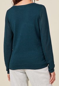 Femme portant un pull en maille turquoise avec des manches longues en mesh et un jean délavé, vue de dos sur un fond neutre.