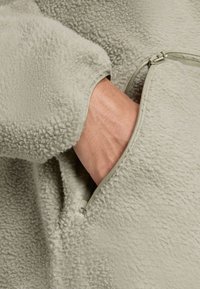 Light green fleece jacket with a soft texture and a zippered pocket, showcasing a close-up of the fabric and hand in the pocket.