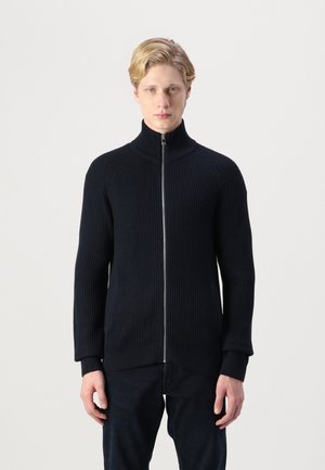 Maglione nero a coste con zip e colletto alto, maniche lunghe e tasche laterali. Il tessuto ha una finitura testurizzata e una silhouette aderente.