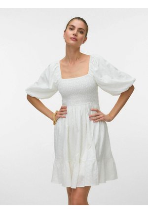 Žena v bielych šatách s nariasenými rukávmi, so smockovaným živôtikom a volánikovým lemom, pózuje s rukami v bokoch na obyčajnom pozadí.