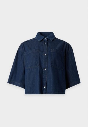 Femme portant une chemise oversize bleu foncé à manches courtes, un pantalon beige à jambes larges, des ballerines noires pointues, tenant un grand sac cabas noir perforé.