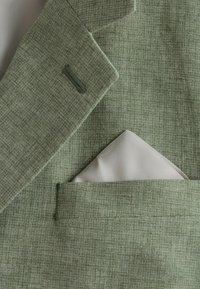Tissu vert texturé d'une veste de costume avec un revers cranté, présentant un mouchoir de poche blanc dépassant de la poche poitrine.