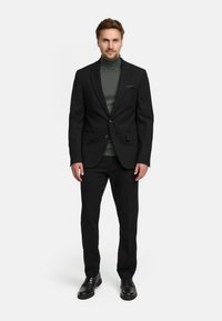 Svart dress med matchende bukser. Båret over en mørkegrønn genser med høy hals. Har en enkelspent design og en lommetørkle. Svarte sko.