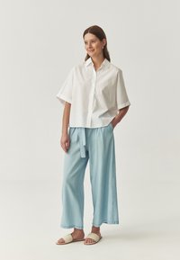 Donna in piedi con le mani in tasca, che indossa una camicia bianca larga con bottoni, pantaloni larghi blu chiaro e sandali beige.