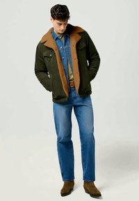 Chaqueta verde con cuello marrón, camisa de mezclilla y jeans azules. La chaqueta tiene un cierre de botones en el frente y dos bolsillos laterales. Botas marrones hasta el tobillo.