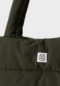 Mørkegrønn polstret stoffveske med et kort håndtak og en liten hvit firkantet etikett med en sirkulær svart logo.