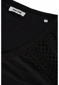 Camiseta negra de cuello en V con tejido texturizado, adornada con intrincados detalles de encaje en un lado y una etiqueta tejida en el cuello.