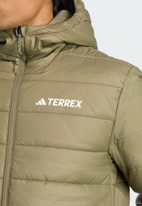 Gros plan sur une personne portant une doudoune olive clair avec capuche, arborant le logo "TERREX" en blanc sur la poitrine.