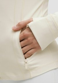 Svijetlo bež atletska jakna s džepom na zip; ima ravne šavove i mekan, rastezljiv materijal. Bliski prikaz ruke i rukava.
