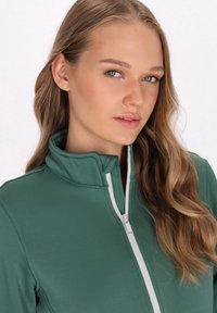 Giacca verde con zip e colletto alto, presentante un tessuto testurizzato e dettagli in zip bianca. Il modello mostra un'espressione neutra.