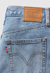 Jean en denim bleu clair avec une patte en cuir portant un texte en relief, des coutures orange contrastantes et une étiquette rouge sur la poche arrière.
