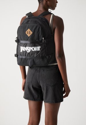 Black Jansport backpack with a textured finish, featuring a brown leather diamond patch and a large white logo at the front. Adjustable straps.