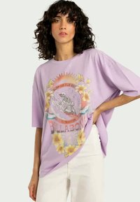 T-shirt roxa de corte solto com um gráfico frontal de um esqueleto a surfar, rodeado por flores e motivos de sol. Mangas curtas, tecido suave.
