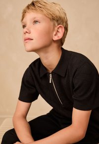 Czarna koszulka polo z krótkimi rękawami i zamkiem przy szyi, z teksturowanym wzorem rombów. Noszona przez młodego chłopca o krótkich blond włosach.