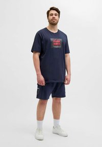 Navy Baumwoll-T-Shirt mit kurzen Ärmeln und grafischem Druck. Kombiniert mit passenden navy Shorts, beide haben einen lässigen Schnitt. Weiße Sneaker runden den Look ab.