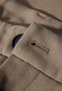 Zbliżenie na beżową tkaninę z czarnym guzikiem częściowo widocznym przez przeszytą dziurkę na guzik na złożonym materiale.