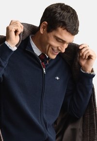 Marineblå genser med glidelås, ribbestrikkede mansjetter, genserstoff, med en liten logo. Modellen legger en brun teksturert frakk over skuldrene.