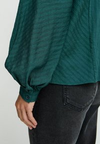 Blusa testurizzata color teal con maniche a palloncino, caratterizzata da un polsino con bottone e strisce diagonali. Abbinata a jeans scuri, vista laterale.