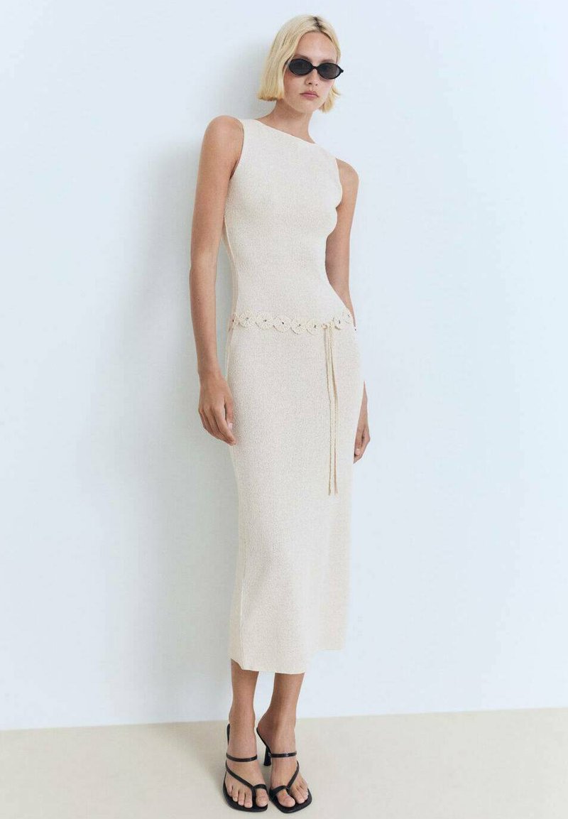Model in een mouwloze, lichtbeige jurk met een decoratieve tailleband en strikjes, gecombineerd met zwarte zonnebril en sandalen met bandjes, staat tegen een bleke achtergrond.