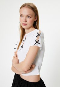 Kurzärmliges weißes Rippenshirt mit schwarzen Schleifenakzenten und Cut-Outs an den Schultern. Gestylt mit einem schwarzen Taillenband darunter.