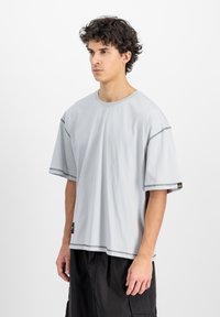 Světle šedé oversized tričko vyrobené z bavlny, s krátkými rukávy, kulatým výstřihem, plochými švy a černou etiketou na boku.