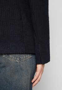 Námornícky texturovaný sveter s žebrovaným designem, s rovnými rukávy a viditelnou rukou opřenou o světle modré džíny.