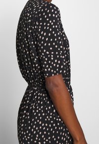 Robe noire à manches courtes, avec un motif à pois beige. Détail de taille élastiquée et texture de tissu douce et fluide.