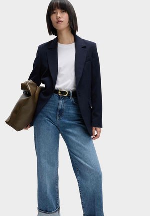 Mujer con blazer negro, camisa blanca, jeans azules con cinturón negro y sosteniendo un gran bolso marrón sobre un fondo liso.