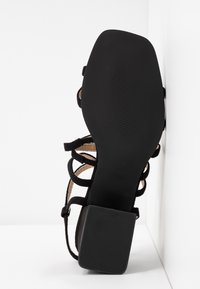 Sandale noire à brides avec un talon carré et une semelle texturée, vue du dessous sur fond blanc.