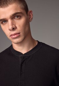Czarny, prążkowany t-shirt henley z okrągłym dekoltem, listwą guzikową i teksturowanym materiałem. Zbliżenie skupiające się na kołnierzyku i detalach szwów.