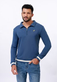 Modrá mikina na zip s límcem, s malým zlatým logem na hrudi, bílými pruhovanými manžetami a žebrovaným lemem. Ležérní design.