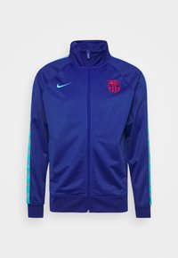 Giacca zip-up blu navy con colletto alto, con logo rosso del Barcellona sul petto e strisce turchesi lungo le maniche. Tessuto liscio.