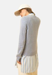 Lichtgrijze gebreide trui met een ronde halslijn, geribbelde zoom en details aan de achternaad, gelaagd over een blauw-wit gestreept shirt.