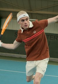 Ruda polo marškiniai su balta apykakle ir raudonomis juostelėmis, derinami su baltais šortais; asmuo judesyje tenisų aikštelėje, laikantis raketę.