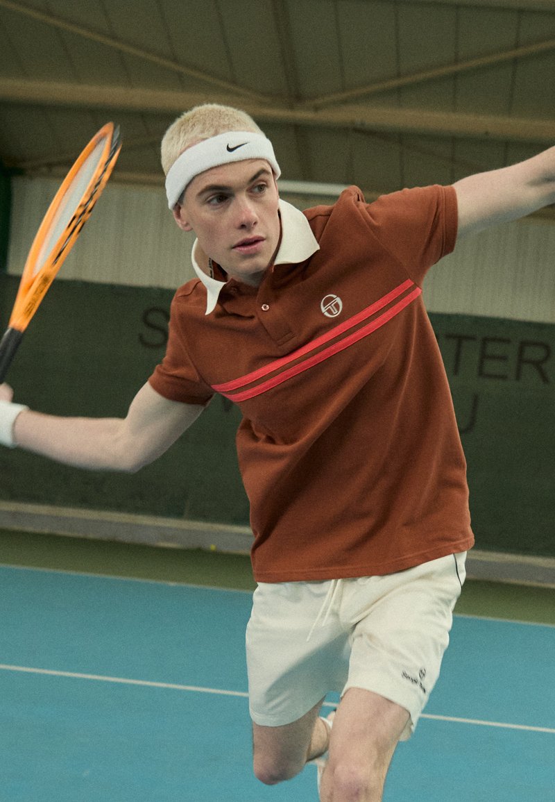 Ruda polo marškiniai su balta apykakle ir raudonomis juostelėmis, derinami su baltais šortais; asmuo judesyje tenisų aikštelėje, laikantis raketę.