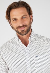 Chemise blanche à boutons avec un petit motif bleu, un col classique et une poche poitrine avec un logo. Texture de tissu lisse.