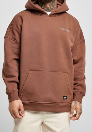 Mann trägt braunen Kapuzenpullover mit Fronttasche und kleinem Logo, kombiniert mit hellbeigen Hosen, zeigt tätowierte Hände und Hals.